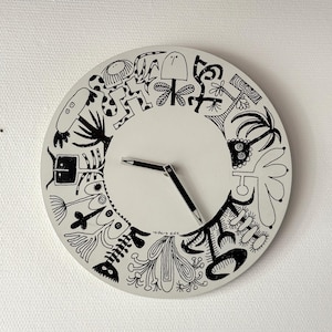 Op de afbeelding: Een ronde wandklok met een lichtgrijze wijzerplaat met een cirkelvormige rand van zwarte inktillustraties van diverse abstracte wezens en planten. De klok heeft twee zwarte wijzers en de handtekening iksen in kleine zwarte letters.