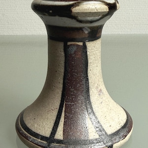 May include: A ceramic candle holder with a flared base and top, featuring a speckled off-white glaze accented by vertical and horizontal dark brown and black painted stripes that create a geometric pattern around the cylindrical body.