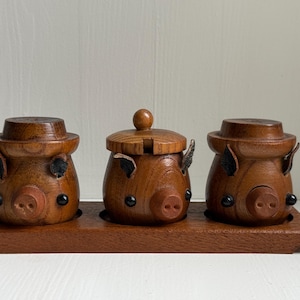 Op de afbeelding: Een houten zout- en peperstel met drie varkensvormige bakjes op een bijpassend houten dienblad. De set is gemaakt van donkerbruin hout en heeft zwarte accenten. Het heeft een rustieke, handgemaakte uitstraling.