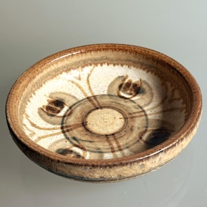 May include: A small, round ceramic dish with a raised rim, featuring a cream-colored interior decorated with brown, hand-painted floral motifs and a central circular pattern. The exterior edge has a textured, mottled brown glaze finish.