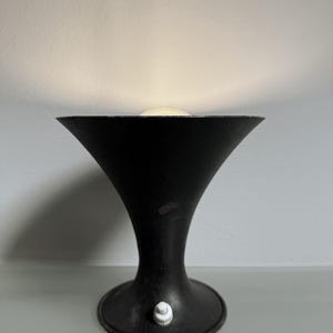 Op de afbeelding: Een zwarte tafellamp in vintage-stijl met een uitlopende kap en een afgeronde voet. De lamp heeft een witte aan/uit-schakelaar en geeft een zacht, warm licht. De lamp is ongeveer 25 cm hoog.