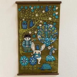 Puede incluir: Un tapiz de pared vintage con una escena forestal caprichosa. La tela verde oliva muestra un árbol con frutos azules, pájaros y un pavo real. También se representan un niño, un conejo, flores y un erizo. El texto "ULLAS." está en la parte inferior.