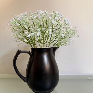 Op de afbeelding: Een zwarte keramische kan met een gebogen handvat, gevuld met een boeket witte en groene gipskruidbloemen. De kan heeft een gladde, matte afwerking en staat op een reflecterend oppervlak.