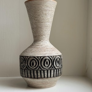 Op de afbeelding: Een hoge, decoratieve vaas met een crèmekleurige body en een gestructureerd, horizontaal gestreept patroon. De vaas heeft een band van zwarte, geometrische ontwerpen in het midden. De rand is warmbruin.