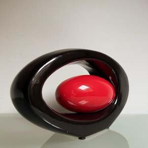 Op de afbeelding: Een moderne, abstracte sculptuur met een glanzend, donkerbruin, ovaalvormig frame met een felrode, eivormige vorm erin. De sculptuur heeft een glad, reflecterend oppervlak en staat op een licht oppervlak.