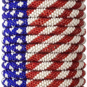 Puede incluir: Una pila de pulseras de cuentas, cada una meticulosamente elaborada con pequeñas cuentas de colores. Las pulseras muestran un diseño patriótico, con rayas rojas y blancas alternas, y un campo azul con estrellas blancas, que se asemeja a la bandera estadounidense.