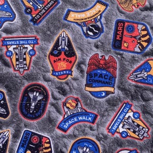 May include: A fabric pattern featuring colorful space-themed patches. The patches include rockets, planets, and phrases like "To the Stars," "Space Walk," and "Aim for the Stars." The background is a textured gray, resembling a lunar surface.