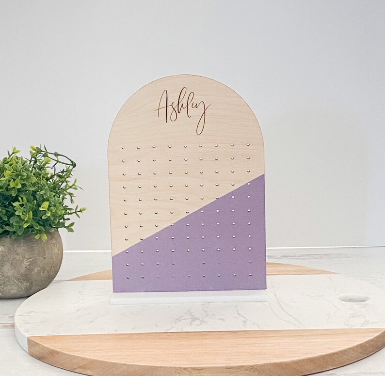 Arch-shaped earring stand engraved with a girl's name and stands on a display block. Lavender is painted across the earring stand that can be customized with your favorite color.