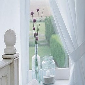 May include: A serene window scene featuring a white bed, curtains, and a windowsill display. A clear glass bottle holds purple flowers, next to a jar with a candle. The window overlooks a green garden.
