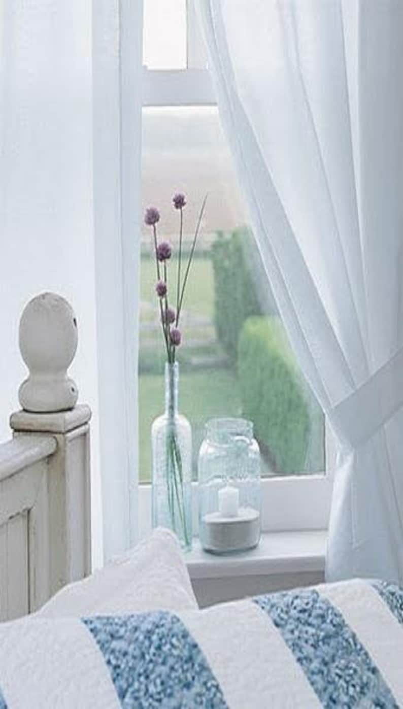 May include: A serene window scene featuring a white bed, curtains, and a windowsill display. A clear glass bottle holds purple flowers, next to a jar with a candle. The window overlooks a green garden.