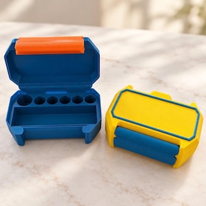 May include: Two small rectangular storage cases. One case is open, featuring a dark blue interior with six circular indentations and a rectangular compartment, topped with an orange hinge. The second case is closed, colored yellow with a blue border.