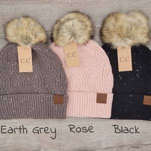May include: Three knitted winter hats with faux fur pom poms. The hats are in earth grey, rose, and black. Each hat has a small brown leather patch with the letters "C.C." on it.