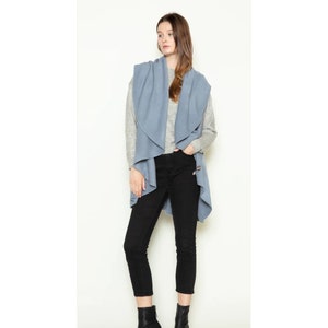 May include: A light blue open-front cardigan vest with a draped collar. The vest is worn over a gray long-sleeve shirt and black pants.