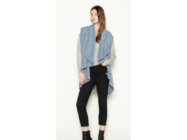 May include: A light blue open-front cardigan vest with a draped collar. The vest is worn over a gray long-sleeve shirt and black pants.