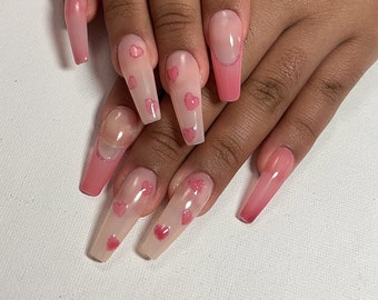 May include: A set of long, coffin-shaped acrylic nails. The nails feature a gradient pink color, transitioning from a nude base to a vibrant pink tip. Some nails are adorned with small pink heart designs and glitter accents, creating a romantic aesthetic.