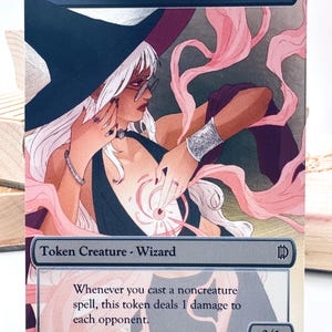 May include: A rectangular trading card titled WIZARD featuring an illustration of a white-haired figure in a black hat and dark clothing, surrounded by pink magical energy. The card text reads: Whenever you cast a noncreature spell, this token deals 1 damage to each opponent. The card includes a 0/1 power and toughness indicator.