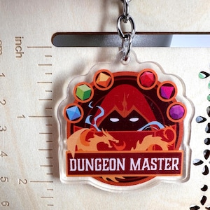 May include: A clear acrylic keychain featuring a hooded figure, a dragon, and six colorful polyhedral dice. The text "DUNGEON MASTER" is printed in white on a red banner. A ruler shows the keychain is approximately 2.5 inches in height.