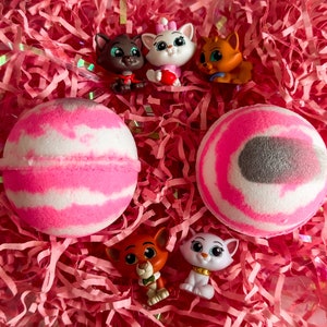 Könnte beinhalten: Zwei rosa und weiße Badebomben mit einem grauen Wirbelmuster, umgeben von rosa Knisterpapier und kleinen Comic-Katzenfiguren.