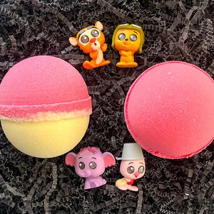 Könnte beinhalten: Zwei rosa Badebomben mit einer gelben und weißen Badebombe. Die Badebomben sind von schwarzem zerkleinertem Papier umgeben. Auf dem zerkleinerten Papier befinden sich vier kleine Comicfiguren von Tieren.