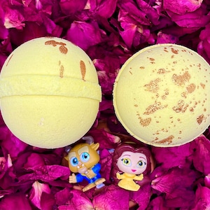 May include: Two yellow bath bombs with gold flecks, a small plastic figurine of Belle from Beauty and the Beast, and a small plastic figurine of the Beast from Beauty and the Beast. The bath bombs and figurines are on a bed of pink rose petals.
