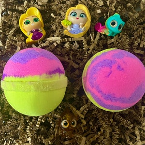 May include: Two yellow and pink bath bombs with a purple swirl design. The bath bombs are surrounded by brown crinkle paper and small plastic figurines of a princess and a chameleon.