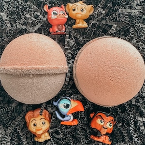 Puede incluir: Dos bombas de baño grandes, redondas y de color marrón claro con una superficie texturizada. Seis pequeñas figuras de personajes de El Rey León están esparcidas alrededor de las bombas de baño. El fondo es papel triturado negro.