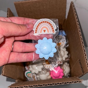May include: A cardboard box filled with small, flower-shaped bath bombs in various colors, including blue, white, and pink. One bath bomb is held in a clear plastic bag with a sticker that says "Thank you for shopping small."