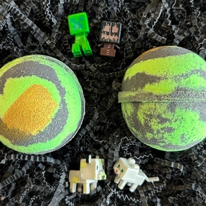 以下が含まれることがあります： 黒のクシャクシャの紙と小さなマインクラフトのキャラクターのフィギュアに囲まれた、緑と黒の2つの金色のキラキラの入ったバスボム
