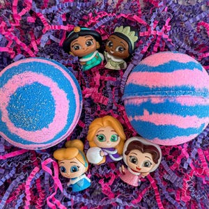 May include: Two large bath bombs with blue and pink stripes, and six small character figurines, are nestled in pink and purple paper filler. The bath bombs have a glittery finish. The figurines are of cartoon characters.