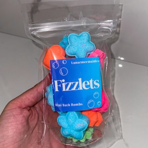 Könnte beinhalten: Ein klarer Plastikbeutel gefüllt mit bunten Mini-Badebomben. Die Badebomben haben verschiedene Formen, darunter Sterne und Herzen, und sind in den Farbtönen Blau, Orange, Pink und Grün gehalten. Ein blaues Etikett trägt die Aufschrift "Fizzlets Mini Bath Bombs".