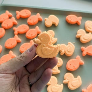 May include: A collection of small, orange, molded bath bombs shaped like ducks and fish. One duck-shaped bath bomb is held up, showcasing its textured surface and distinct outline, while others are scattered across a light green surface.