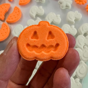 Puede incluir: Una bomba de baño de color naranja brillante con forma de calabaza y un rostro de linterna tallado con ojos triangulares y boca ondulada. La superficie tiene un acabado de polvo comprimido texturizado y el objeto es de perfil circular.