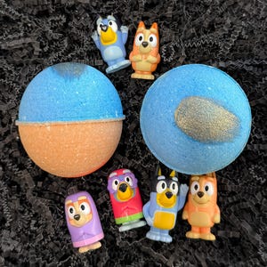 May include: A top-down view of two blue and orange bath bombs with gold glitter accents, surrounded by small character figurines. The figurines are various colors and depict cartoon characters. The background is black.