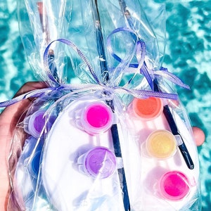 May include: Paint palettes with various colors of paint and paintbrushes are packaged in clear cellophane and tied with a purple ribbon. The palettes are white and shaped like an egg.