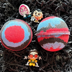 May include: Two round bath bombs with red, blue, and black concentric circles, and three small character figurines on a bed of black shredded paper. The bath bombs have a textured, granular surface.