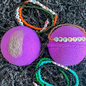May include: Two purple bath bombs with beaded bracelets. One bath bomb has a silver glitter accent. The bracelets spell out "LOVE" and "SWIFTIE" with colourful beads. Additional bracelets in green, black, and white are also visible.