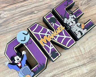 Puede incluir: Letras "UNO" moradas y negras con un contorno dorado. Las letras están decoradas con una telaraña, murciélagos y personajes de Mickey Mouse vestidos de vampiro y momia. Las letras también incluyen el nombre "Manuel".