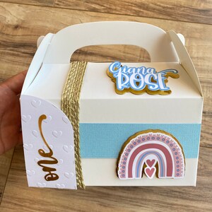 K&ouml;nnte beinhalten: Eine wei&szlig;e Geschenkbox mit Griff, verziert mit einem blauen und rosa Regenbogen, zwei Herzen und einem goldenen Etikett mit der Aufschrift "one". Die Box hat auch ein goldenes Etikett mit dem Namen "Giana Rose" in blauen und wei&szlig;en Buchstaben.