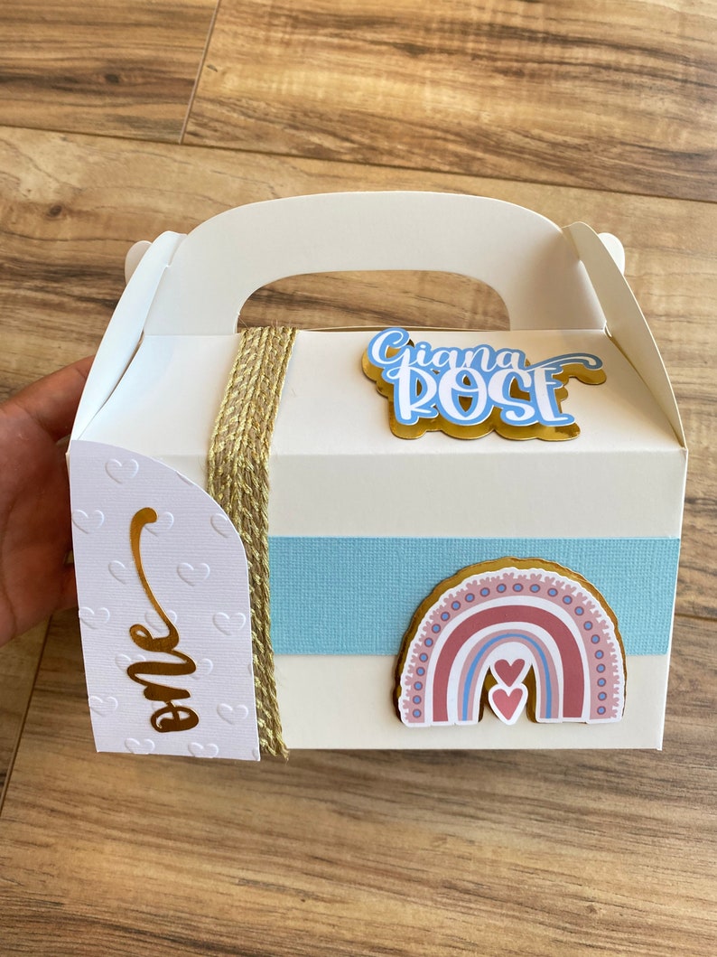 K&ouml;nnte beinhalten: Eine wei&szlig;e Geschenkbox mit Griff, verziert mit einem blauen und rosa Regenbogen, zwei Herzen und einem goldenen Etikett mit der Aufschrift "one". Die Box hat auch ein goldenes Etikett mit dem Namen "Giana Rose" in blauen und wei&szlig;en Buchstaben.