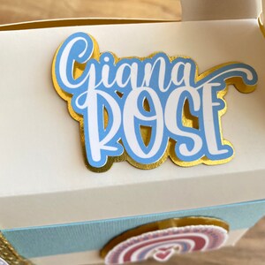 K&ouml;nnte beinhalten: Eine wei&szlig;e Geschenkbox mit einem blauen und goldenen Dekoaufkleber mit der Aufschrift "Gianna Rose" und einem Regenbogenaufkleber mit einem Herz.