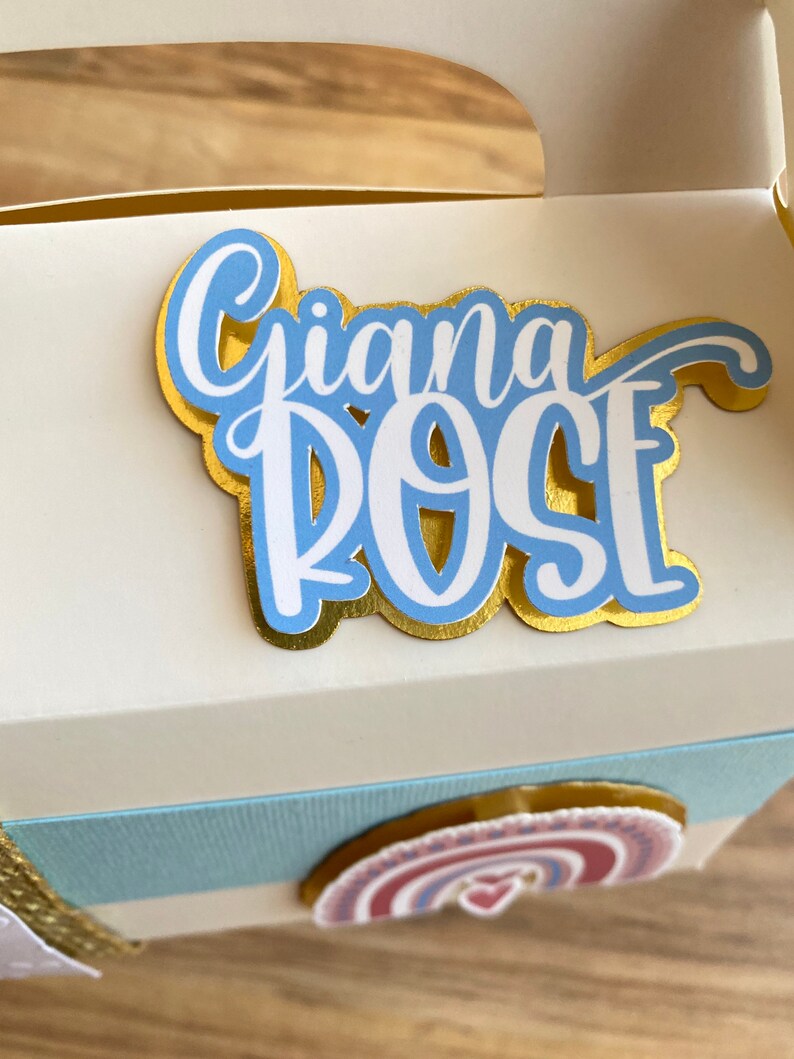 K&ouml;nnte beinhalten: Eine wei&szlig;e Geschenkbox mit einem blauen und goldenen Dekoaufkleber mit der Aufschrift "Gianna Rose" und einem Regenbogenaufkleber mit einem Herz.