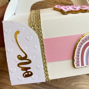 K&ouml;nnte beinhalten: Wei&szlig;e Geschenkbox mit goldener Kordelverzierung, rosa Schleife und einer goldenen Folie "eins" an der Seite. Die Box hat einen Griff und einen Regenbogenaufkleber auf der Vorderseite.