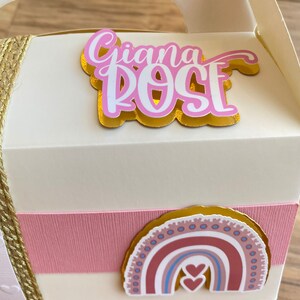 K&ouml;nnte beinhalten: Eine wei&szlig;e Geschenkbox mit einem rosa und goldenen Band. Die Box hat einen Aufkleber mit dem Namen "Giana Rose" und einen Regenbogen-Aufkleber mit zwei Herzen.