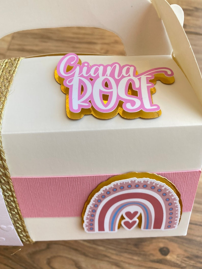 K&ouml;nnte beinhalten: Eine wei&szlig;e Geschenkbox mit einem rosa und goldenen Band. Die Box hat einen Aufkleber mit dem Namen "Giana Rose" und einen Regenbogen-Aufkleber mit zwei Herzen.