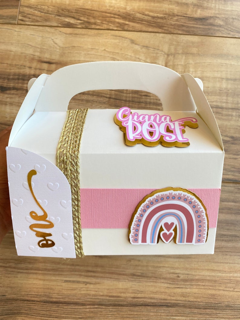 K&ouml;nnte beinhalten: Eine wei&szlig;e Geschenkbox mit goldenem Griff und einem rosa-goldenen Band. Die Box hat einen rosa-goldenen Regenbogen-Aufkleber mit zwei Herzen und dem Text "Giana Rose" in rosa-goldenen Buchstaben. Die Box hat auch einen wei&szlig;en Aufkleber mit dem Wort "one" in goldenen Buchstaben.