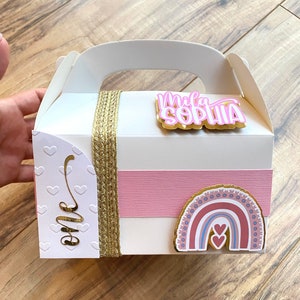 K&ouml;nnte beinhalten: Wei&szlig;e Geschenkbox mit goldener Schleife und einem rosa-goldenen Regenbogen-Aufkleber. Die Box hat ein rosa-wei&szlig; gestreiftes Band und einen Aufkleber mit der Aufschrift "Mila Sophia" in rosa Buchstaben auf goldenem Hintergrund. Die Box hat auch einen Aufkleber mit der Aufschrift "one" in goldenen Buchstaben auf wei&szlig;em Hintergrund.
