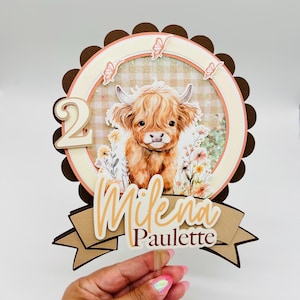 May include: A brown and white Highland cow cake topper with a floral background. The topper features the number "2" and the name "Milena Paulette".