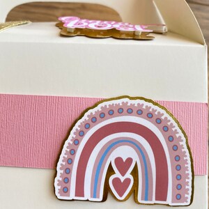 K&ouml;nnte beinhalten: Eine wei&szlig;e Geschenkbox mit einem rosa Band und einem goldenen Regenbogenaufkleber mit zwei Herzen. Der Regenbogen ist in den Farben Rosa, Braun, Blau und Wei&szlig; gehalten.