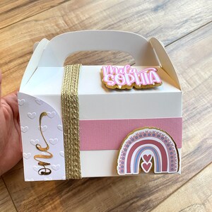 K&ouml;nnte beinhalten: Wei&szlig;e Geschenkbox mit einem rosa-goldenen Regenbogen-Aufkleber, einem rosa-goldenen "Mila Sophia"-Aufkleber und einem goldenen "Eins"-Aufkleber. Die Box hat eine braune Kordelschleife und ein rosa Band.