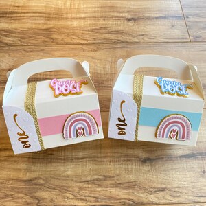 K&ouml;nnte beinhalten: Zwei wei&szlig;e Geschenkboxen mit goldenen Griffen und einem rosa und blauen Band. Die Boxen haben ein Regenbogen-Design mit Herzen und den Text "One" und "Giana Rose".